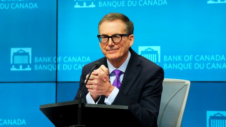 A man in a blue suit, purple tie and glasses is seen sitting at a small podium with a microphone.