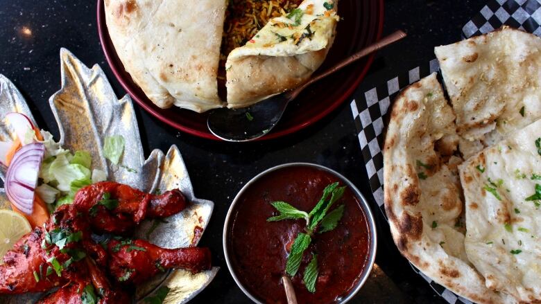 A overhead shot shows a spread of Indian food: bright red, coated drumsticks, a deep red saucy dish, a plate of flatbread, and a bowl covered in a thin bread that has been sliced to reveal yellow rice underneath.