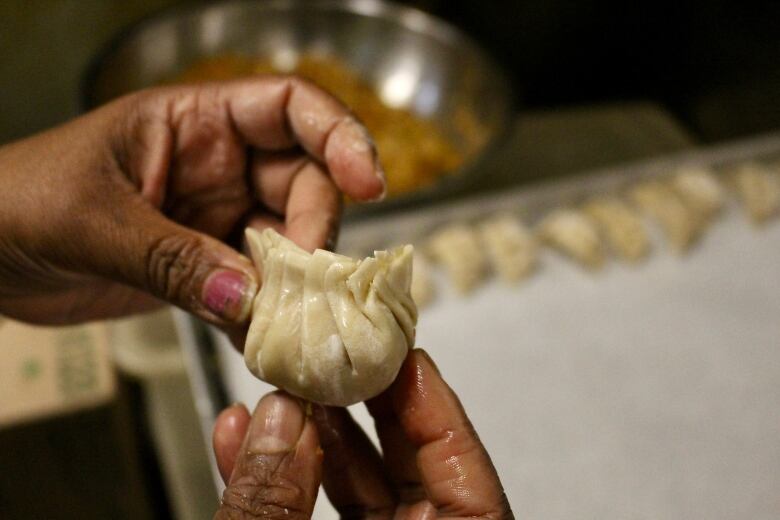 Hands pinch a dumpling together. In the background, you can see a tray with a row of them already complete.