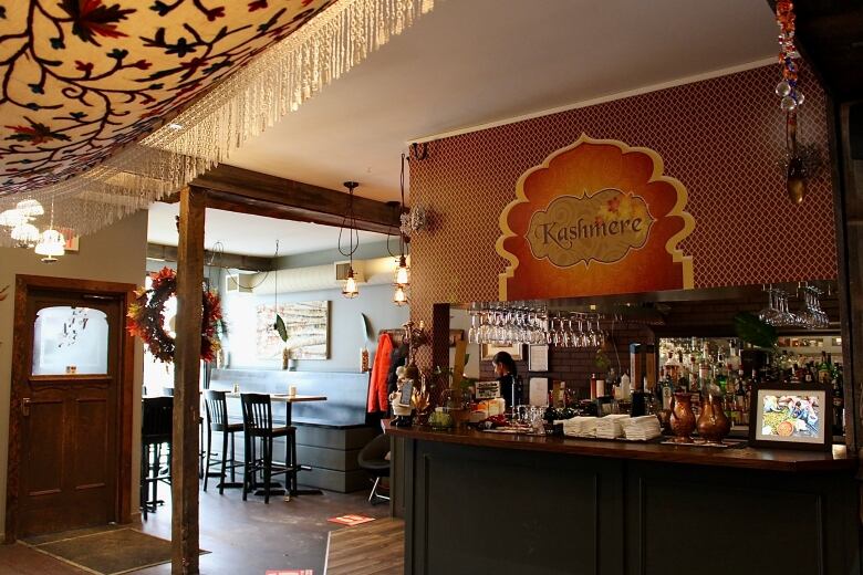 The interior of a restaurant decorated with warm woods; a fringed, cream-coloured scarf; hanging, exposed lightbulbs; and a packed bar topped with an ornately decorated orange sign reading 'Kashmere.'