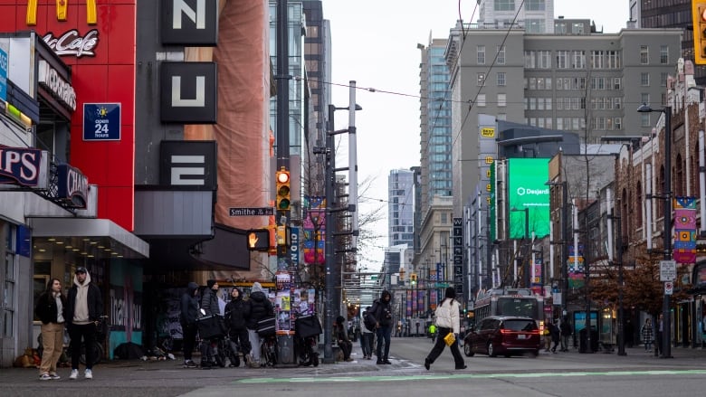 People congregate, cars go by and people cross the street on a busy city road.