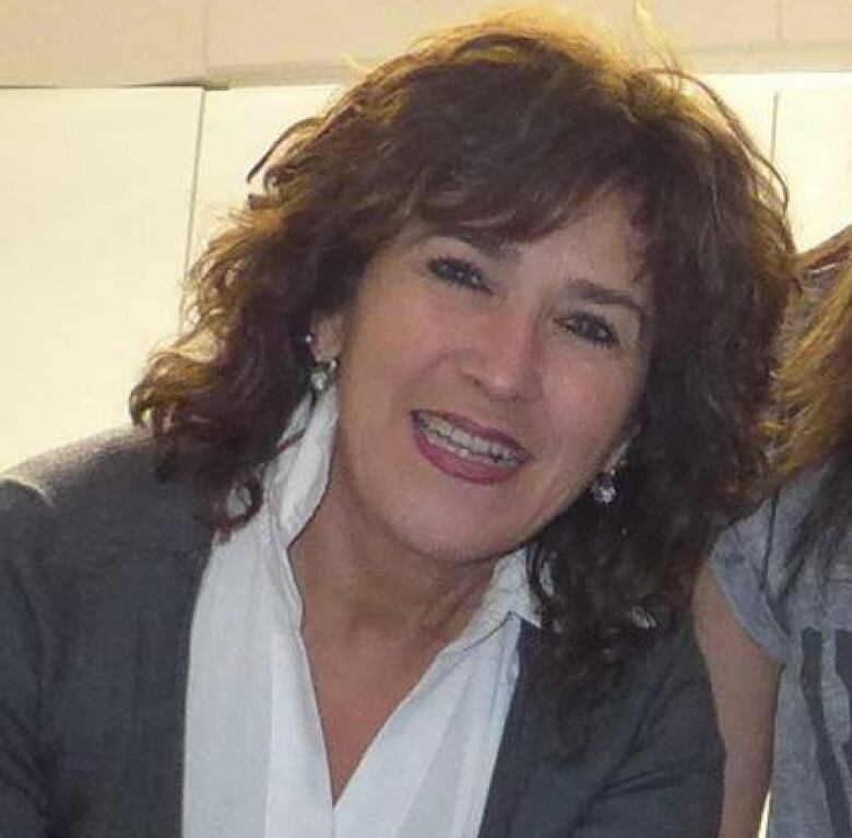 A smiling white woman with shaggy dark brown hair, a white shirt and a grey blazer is shown leaning against another person, who has been cut from the photo.