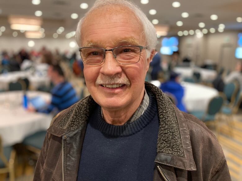 An older man, with receding white hair and glasses, is smiling.