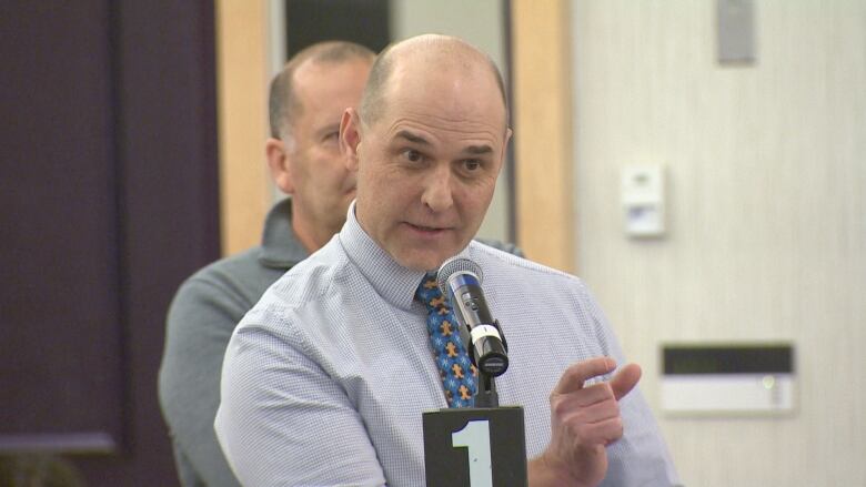 A balding man in a blue shirt and tie stands at a microphone, speaking.