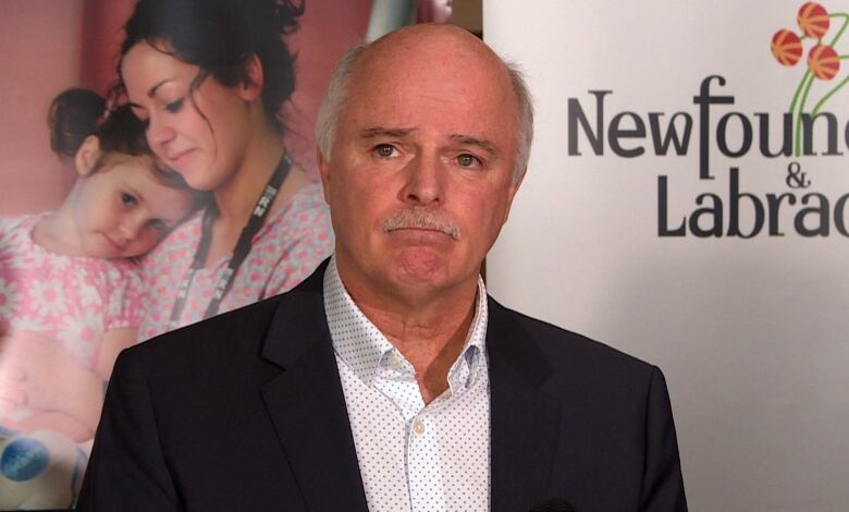 A man wearing a black coat and a white shirt standing in front of a banner which reads Newfoundland and Labrador. 