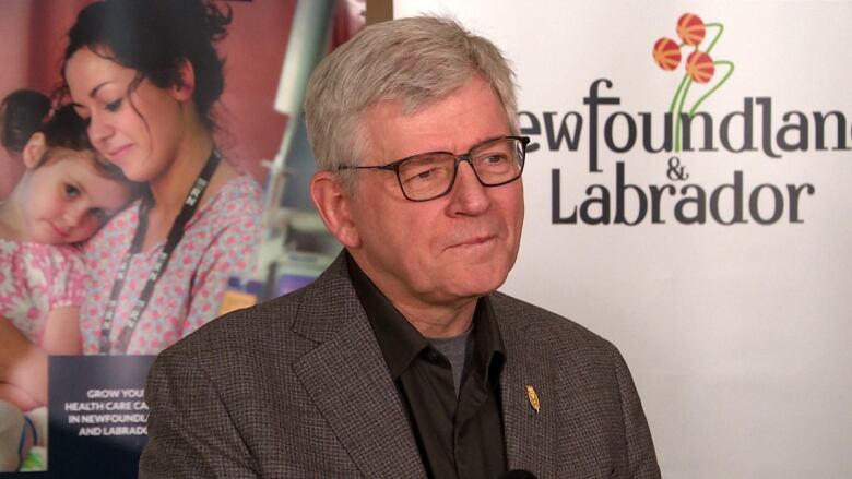 A man with glasses wearing a grey suit jacket and a black shirt standing in front of a banner which reads Newfoundland and Labrador. 