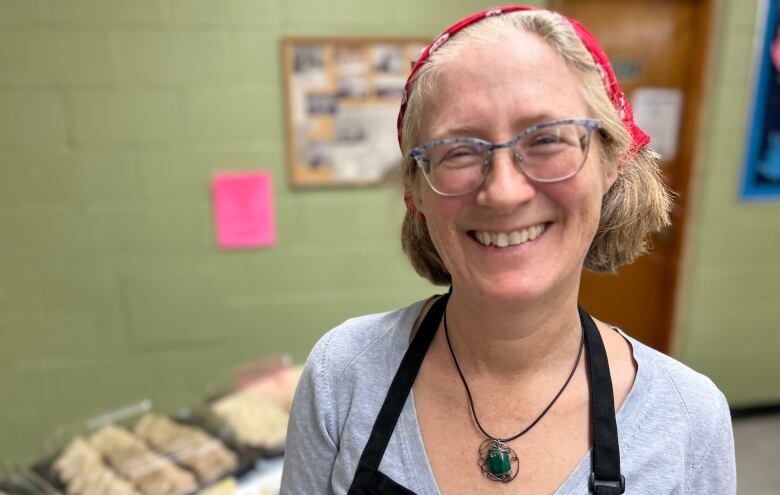 A woman in glasses wearing an apron and a red bandana smiles. A table with a spread of sandwich trays can be seen in the background.