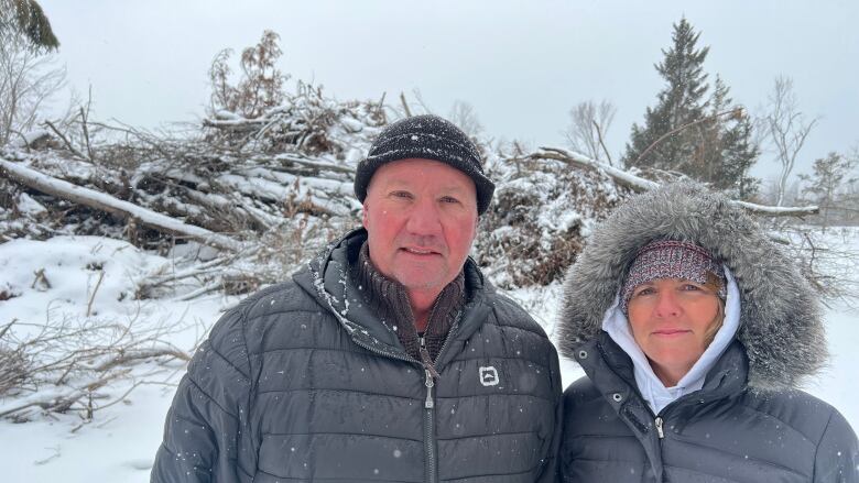 A man and woman in black winter coats stare through heavy snow toward the camera. The pile of tree trunks behind them is covered by snow.