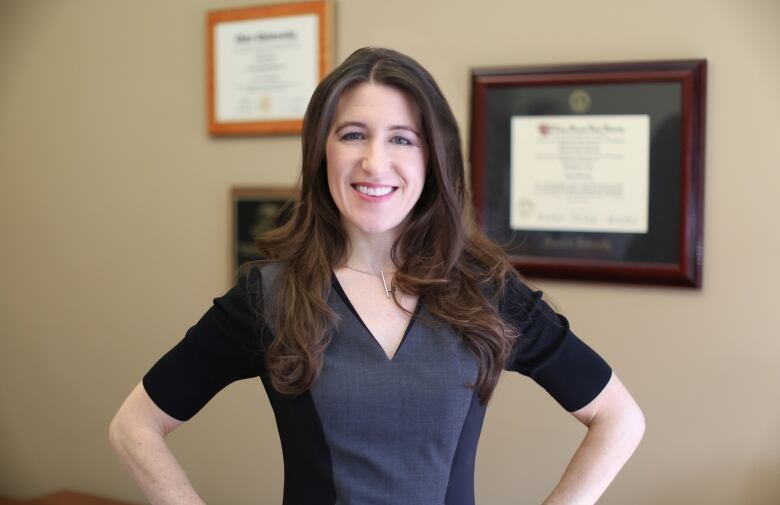 A woman poses with hands on her hips in an office with degrees framed on the wall.