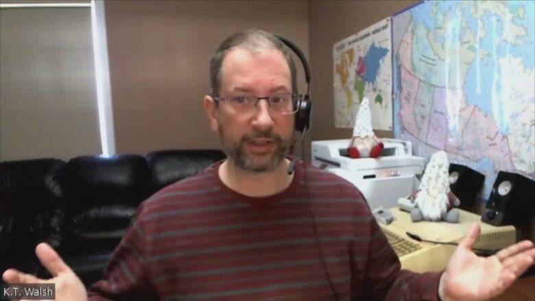 A still from a Zoom. Walsh wears a red and grey striped shirt, glasses and a headset. On his office wall is a map of Canada and the world. 