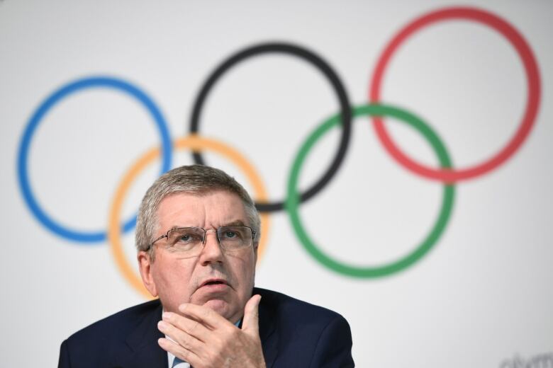 A man in a suit sits in front of a wall depicting the Olympic rings.