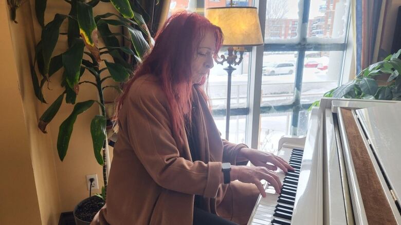 A woman with long red hair playing the piano in front of a window.