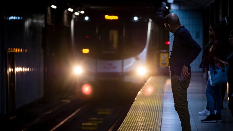 A dimly-lit image of a person on a subway platform looking towards an oncoming train.