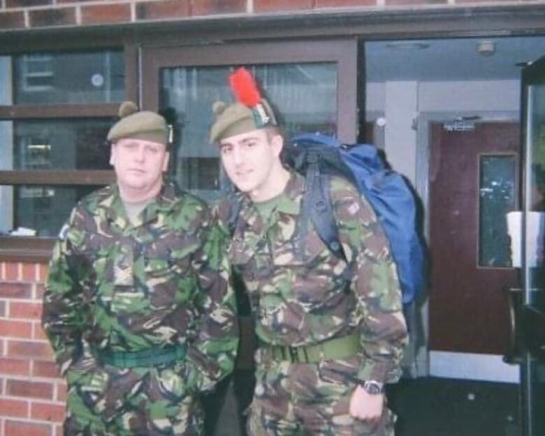 Two men in green camouflage stand in front of a building.