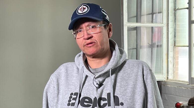 A man with glasses, a hat and grey sweater stands near an open window.