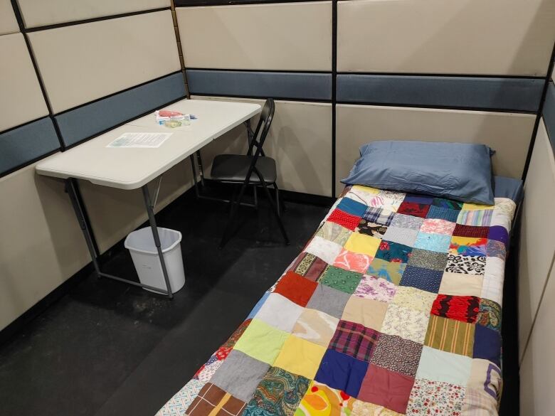 In the right corner of a tall cubicle sits a cot with a colourful quilt and pillow with a blue pillow case. To the left is a foldable table, a foldable chair. A small white garbage bin is beneath the table. On the table, sits a piece of paper and plastic bag filled with toiletries.