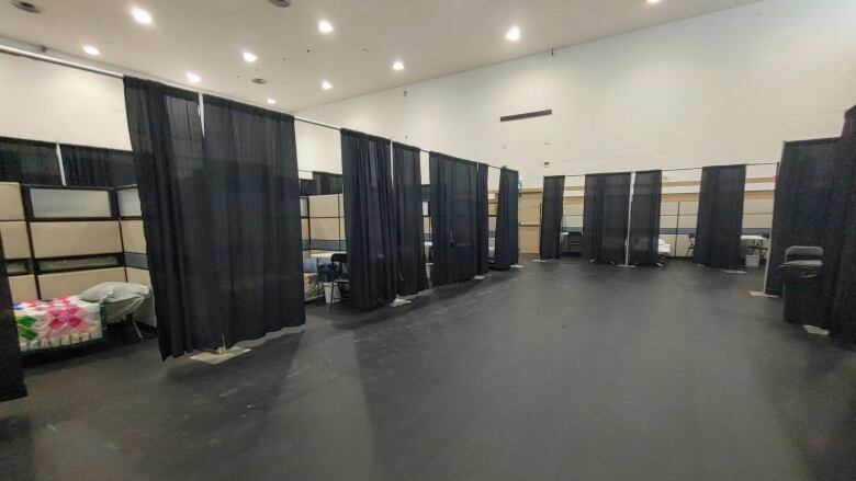 Tall cubicles, with black curtains hanging in front of their entrances, are packed inside an old gymnasium. Inside the cubicles are cots and desks.