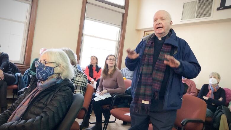 A priest stands and speaks in a room full of seated people.