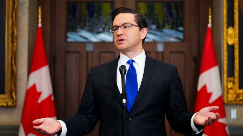 A man in a suit gestures while speaking, flanked on both sides by Canadian flags.