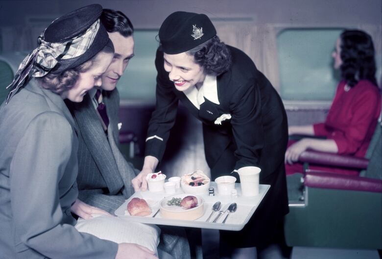 An old-fashioned photo shows a woman serving a couple a meal aboard an airplane