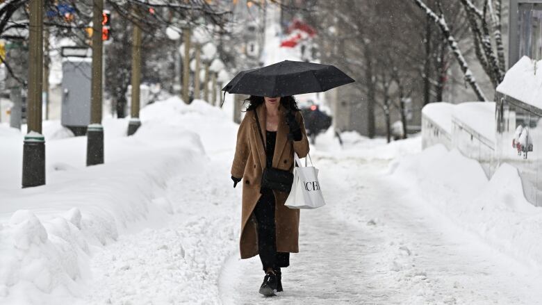 Someone in a brown winter coat walks on a snowy street, holding an umbrella.