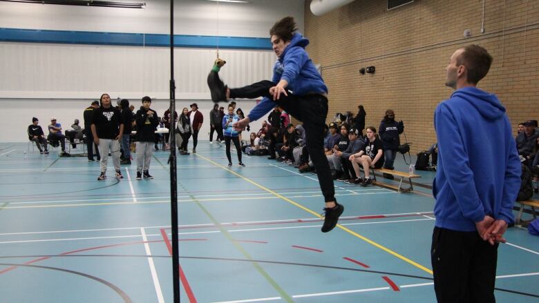 A teenage boy jumps high in the air while performing a kick in a gym while a coach and other athletes watch.