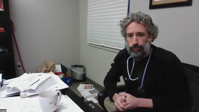 A man sits in front of a desk piled with papers