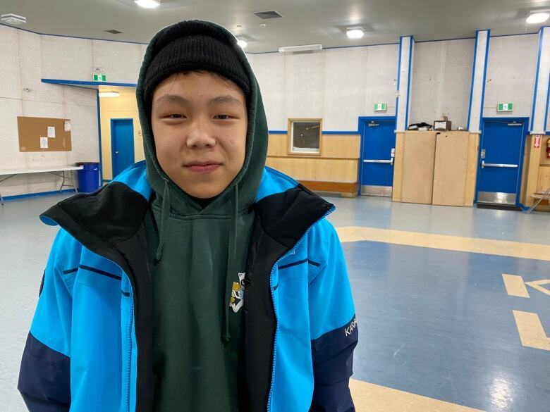 A teenaged boy in a hoodie and jacket stands in an empty gymnasium.