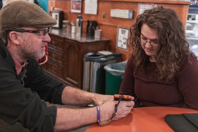 Two people sit at a wooden table in a caf looking at a cellphone with a wooden credenza topped with condiments in the background.