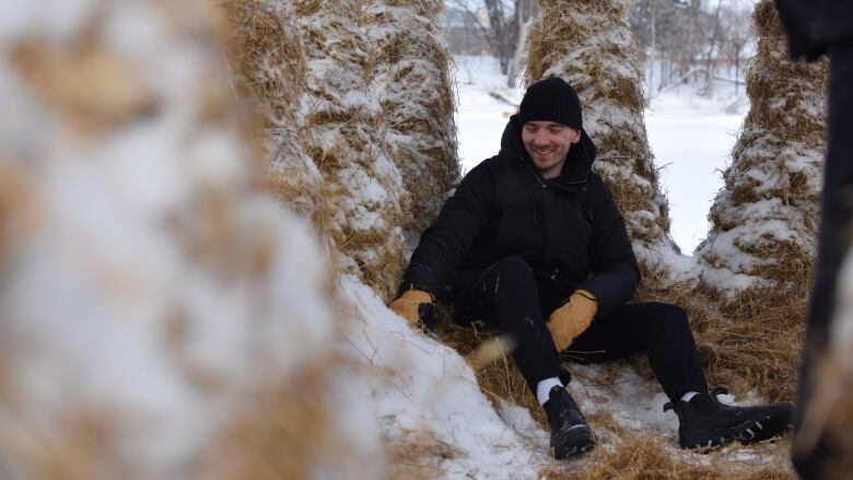 A smiling man, dressed in all black and a black tuque, sits on hay while surrounded by other columns of hay.