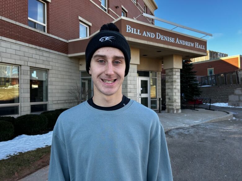 Portrait of a man in a toque and a grey sweater standing in front of a residence hall. 