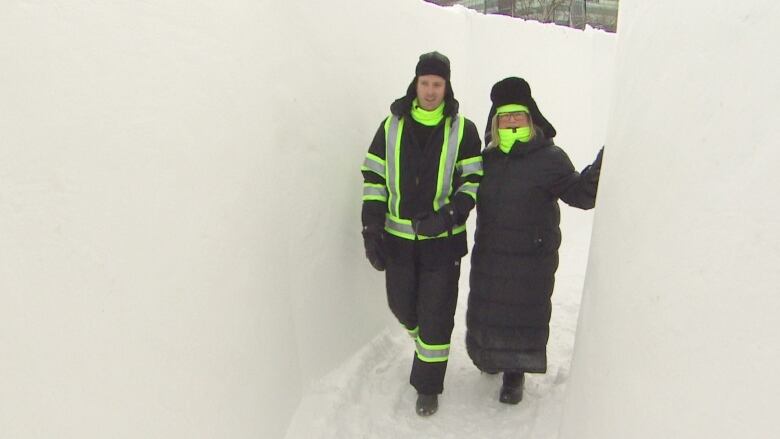 Two people in black winter clothes with bright yellow highlights walk towards the camera, through a hallway of snow.