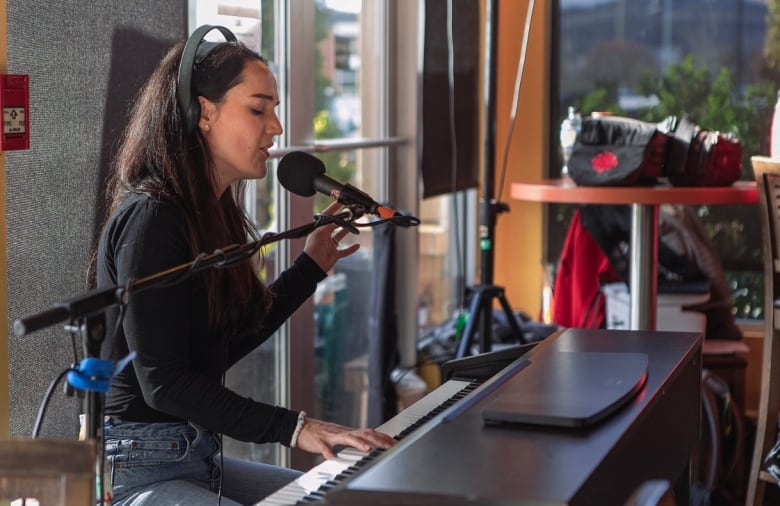 A woman is sitting by the piano wearing a black top and blue jeans, singing into a microphone.