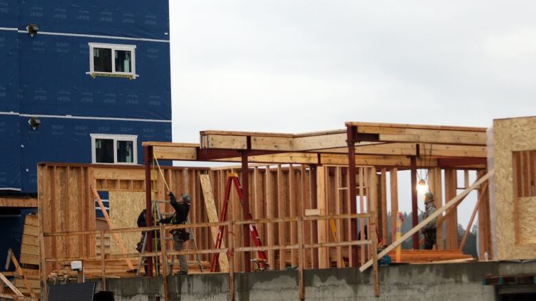 A few builders work on the wooden frames for a home, in the shadow of a large under-construction building with blue tarp on it.