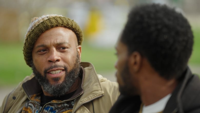 Two black men, one with with a beard smiling, wearing a beige jacket and a knitted had talking on a bench. The second man also has a black beard and hair.