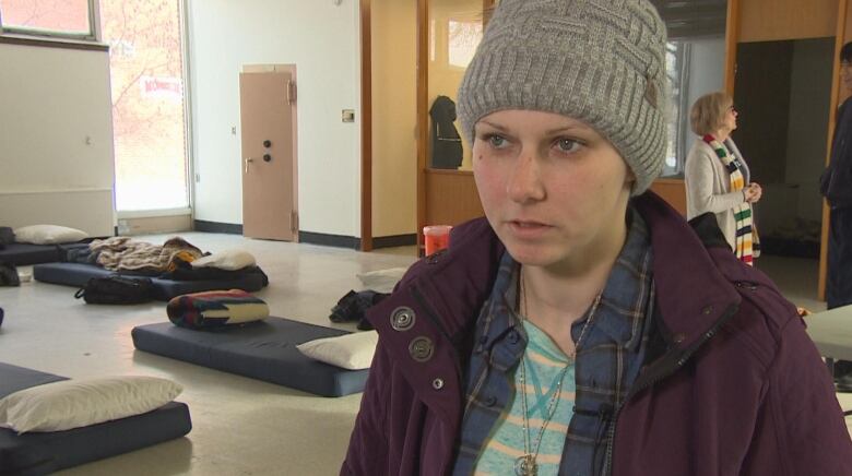 A woman wearing a grey toque, inside a homeless shelter with beds laid on the ground.