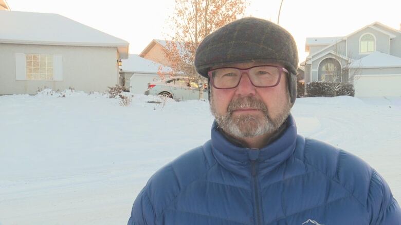 A man stands in front of a house. he is wearing a blue jacket and he has a beard. he is wearing glasses