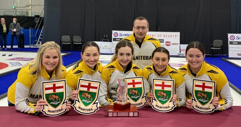 Five female Manitoba curlers pose behind a trophy in front of a male coach.