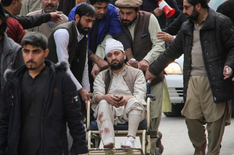 A man with a bandaged head in a wheelchair is wheeled by another man.