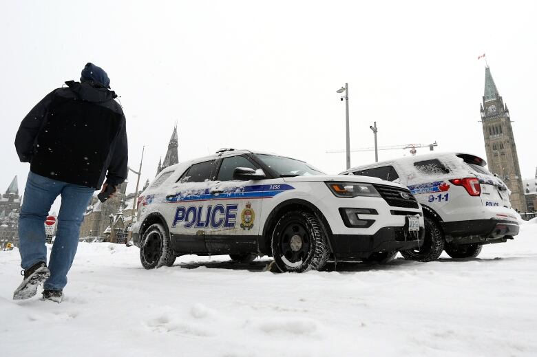 Someone passes two police vehicles near a tall Gothic-style tower in the snow.