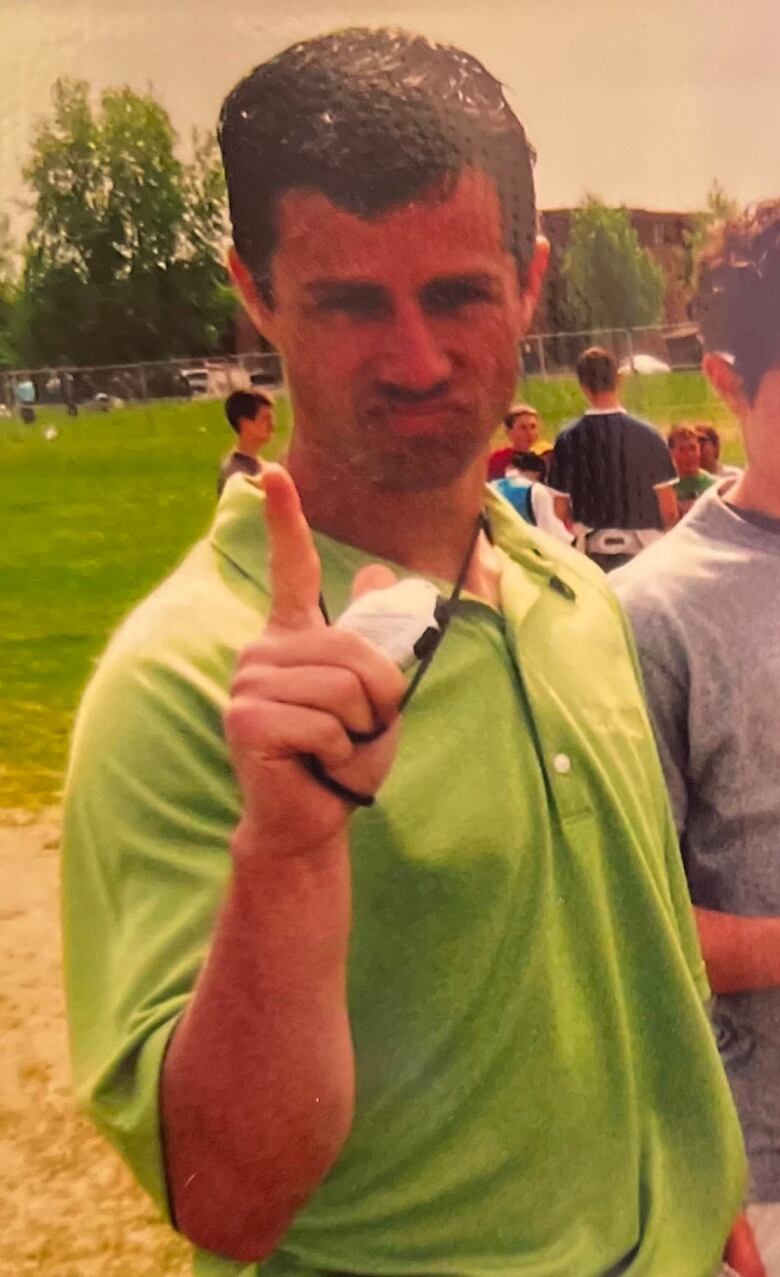 A man in a green shirt is holding up his finger, as if giving a number one sign, to the camera. He is standing outdoors on a sports field.