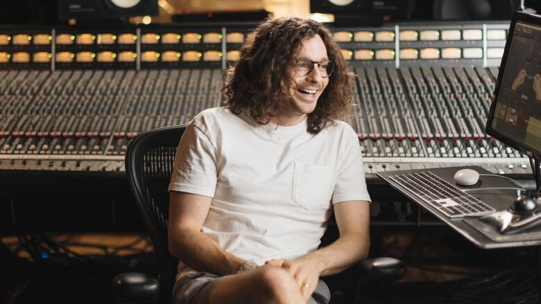 A man in glasses with long curly brown hair sits in a recording studio. He is smiling and wearing a white t-shirt and shorts. 