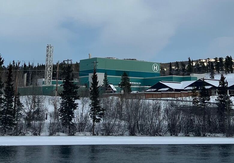 A large green building marked with an 'H' is seen in winter, on the other side of a partially-frozen river.