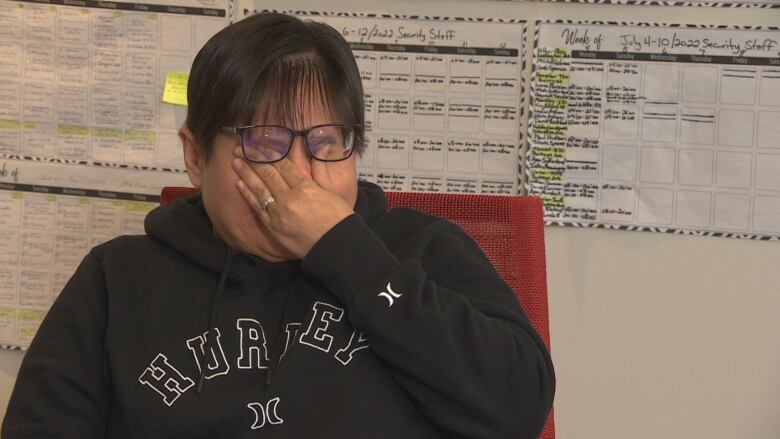 A young woman sitting in a black hoodie with one hand up to her eyes as she cries. 