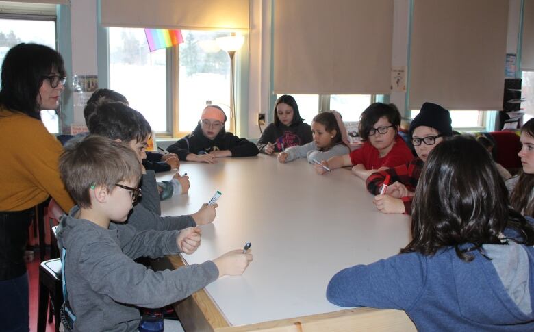 a group of elementary school children sit around a white table with dry erase markers in hand as teacher looks on