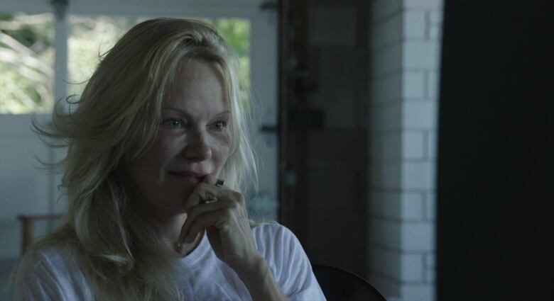 Blonde woman stares at television in dark room in white T-shirt with nail in mouth