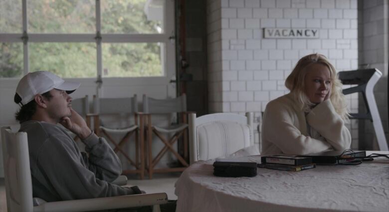 A boy in a grey sweater and white hat and a blonde woman stare into a television watching old VHS tape