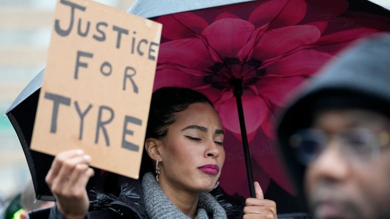 A woman with her eyes closed holds an umbrella and a sign that says 