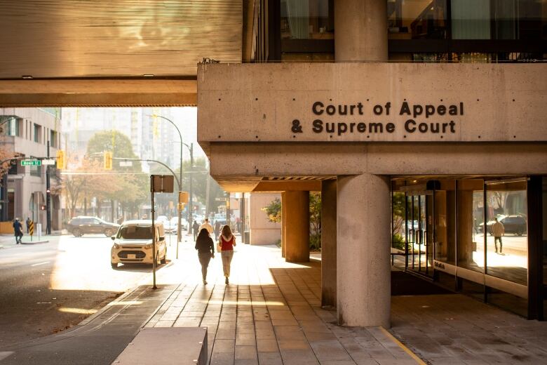 Two people walk down a sunlit street past concrete pillars that support a concrete awning with the words Court of Appeal & Supreme Court over a glass facade.