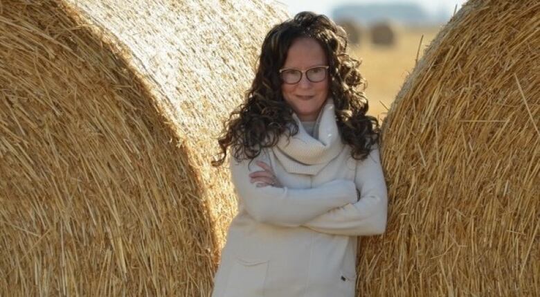 A photo of a woman with curly hair, leaning against a haystack, smiling at the camera.